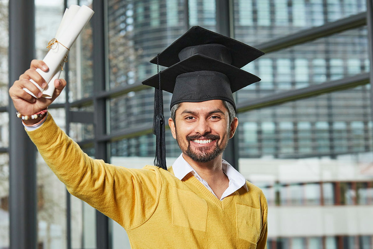 Absolvent mit Graduation Cap hebt Zertifikat in Frontansicht, urbaner Hintergrund.
