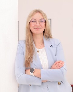 Frau mit langen blonden Haaren in hellblauem Blazer, die selbstbewusst die Arme verschr&auml;nkt.