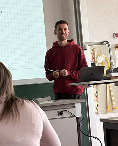 Junger Mann steht in einem Klassenzimmer am Rednerpult, pr&auml;sentiert ein Blatt.
