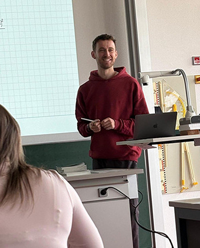Junge Dozent steht hinter Laborpult, präsentiert vor Whiteboard im Klassenzimmer.