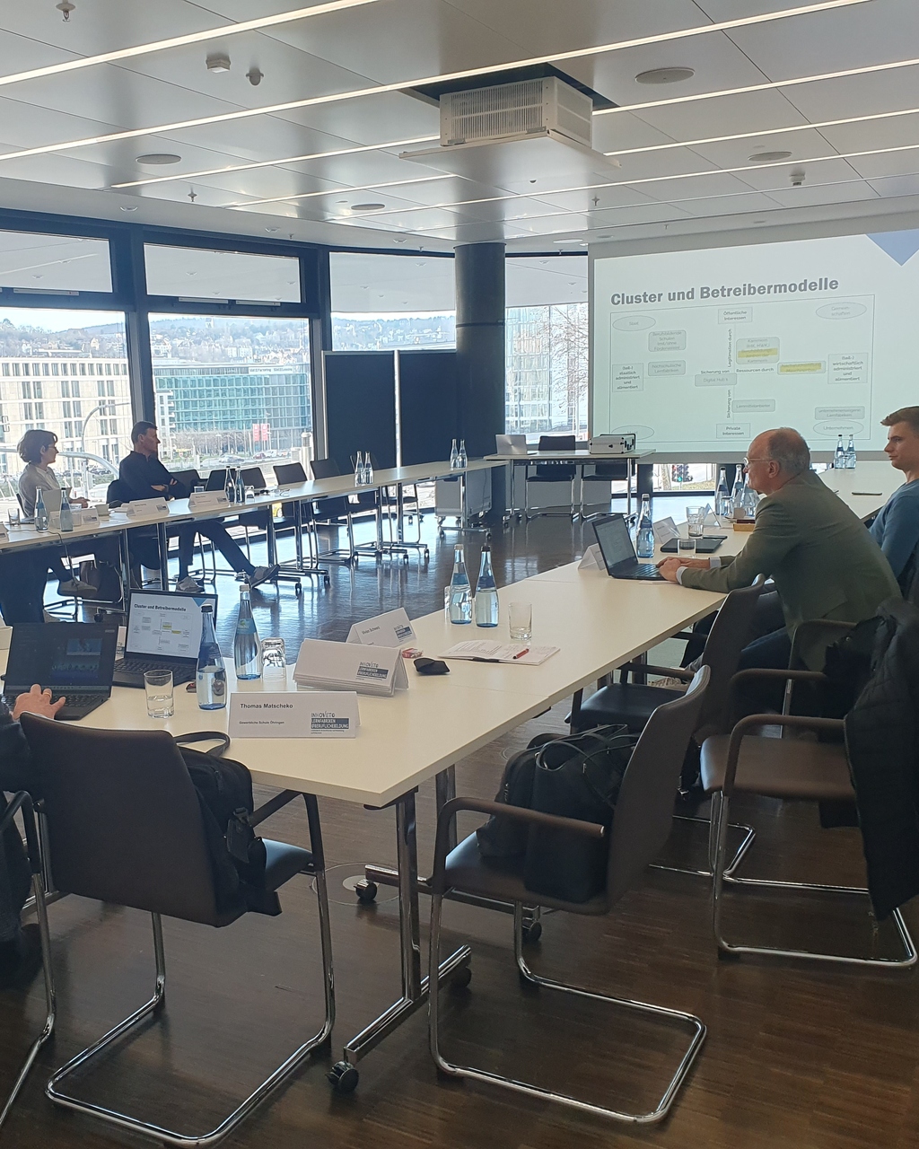 Konferenzraum mit Teilnehmern, Laptops, Wasserflaschen, Tischen und Projektionswand.
