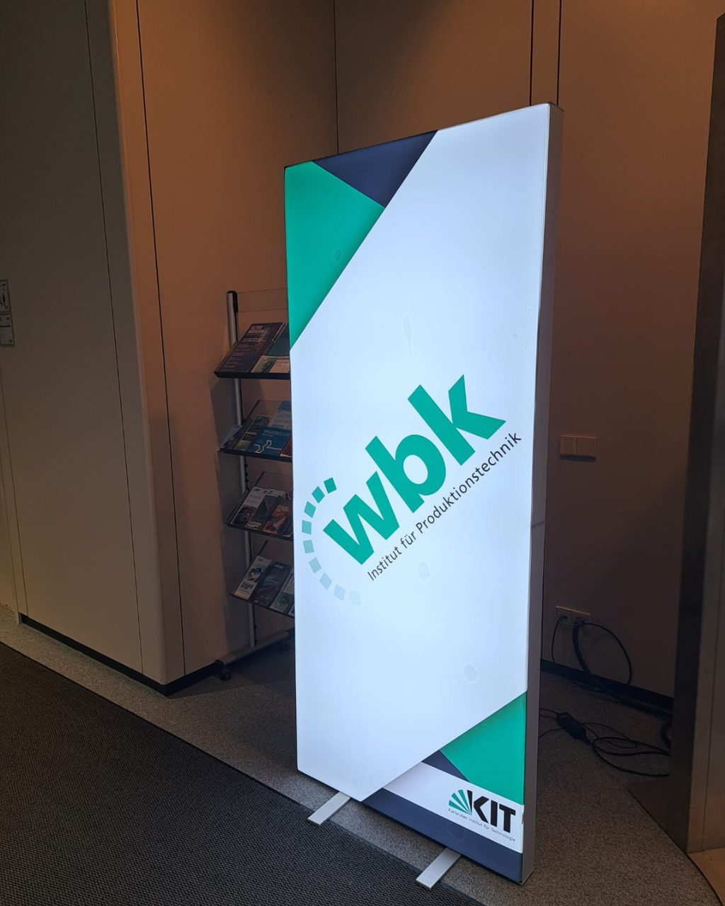 Werbe-Standup mit Logo „ wbk – Institut für Produktionstechnik “ und KIT-Logo, beleuchtet im Innenraum.