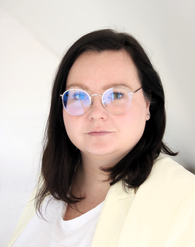 Person mit dunklen Haaren, helle Blazer-Jacke und wei&szlig;es Oberteil vor Innenraum.