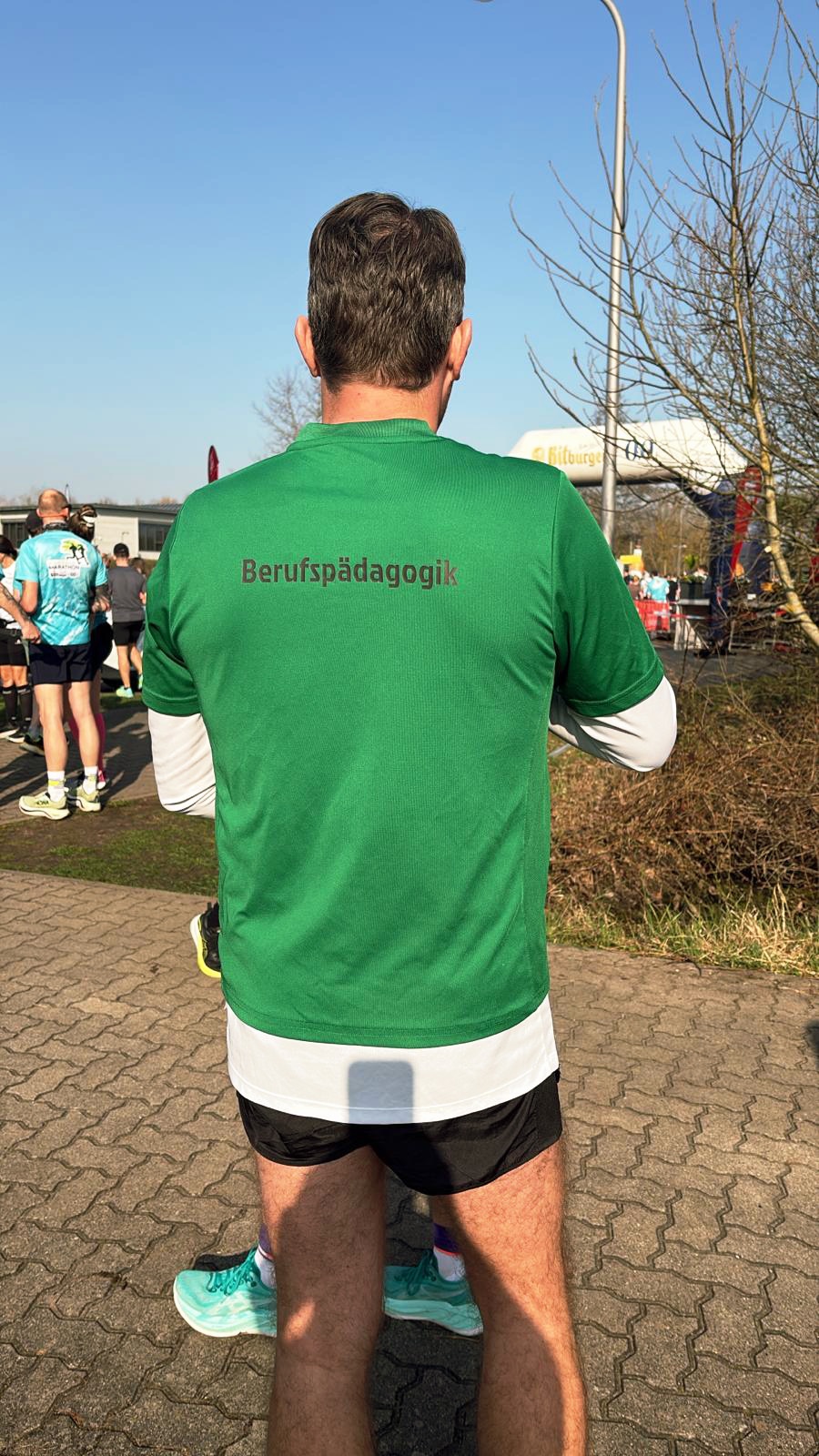Mann von hinten in grüner Laufjacke mit Aufschrift „Berufspädagogik“ bei Outdoorsportveranstaltung.