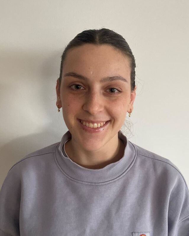 Person mit dunkelhaarigem Haar, seitlich glatte Haare, graues Sweatshirt, neutraler Hintergrund.