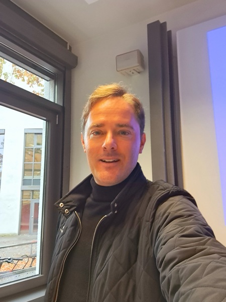 Mann steht in Innenraum neben Fensterfront, Unscharfes Gesicht, schwarze Jacke.