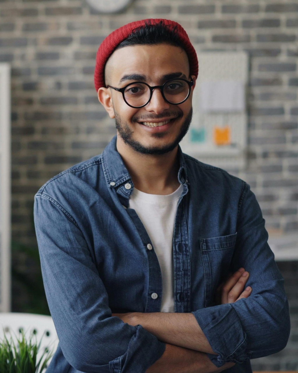Junger Mann mit roter Mütze und Jeanshemd sitzt vor Backsteinwand mit verschränkten Armen.